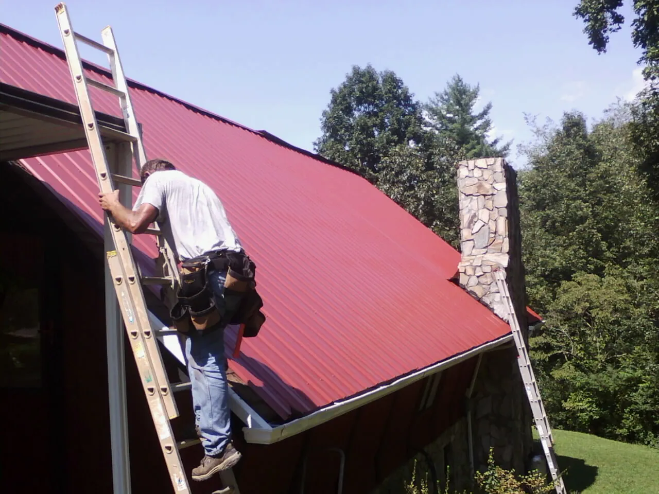 Roofer on a ladder beside a red metal roof and stone chimney — Soffit & Fascia Repair by Roofing Brothers in Connersville